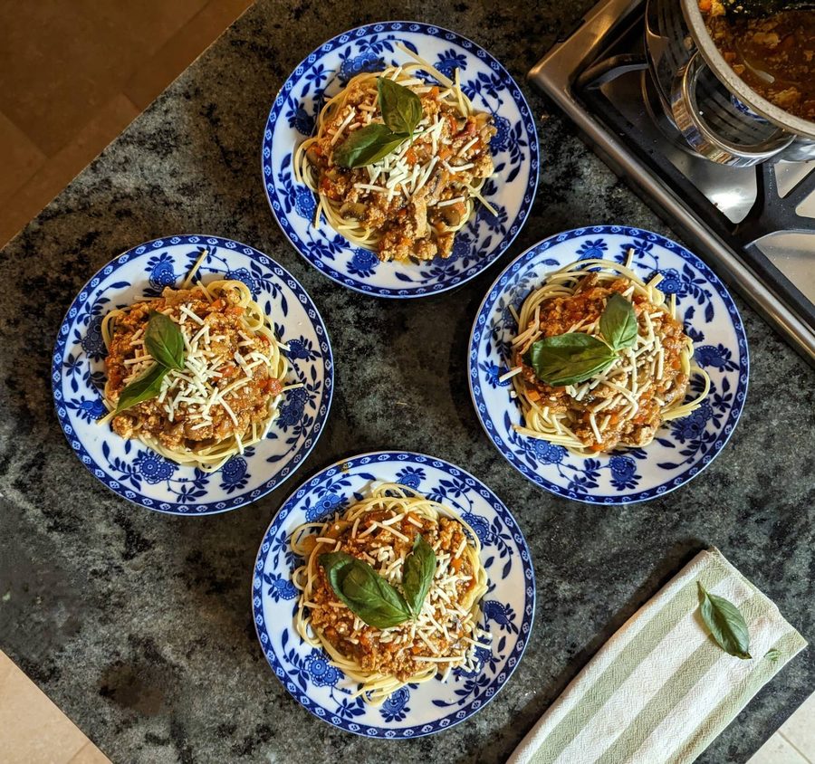 Four blue and white plates of spaghetti topped with vegan bolognese, grated vegan cheese and basil leaves on a kitchen counter.