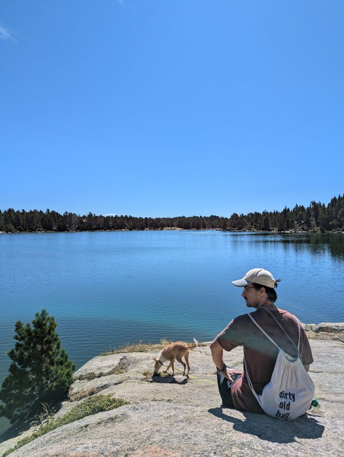 A man in a cap sitting on a granite rock beside a mountain lake under a clear blue sky, with a small rust and white dog sniffing the ground next to him.