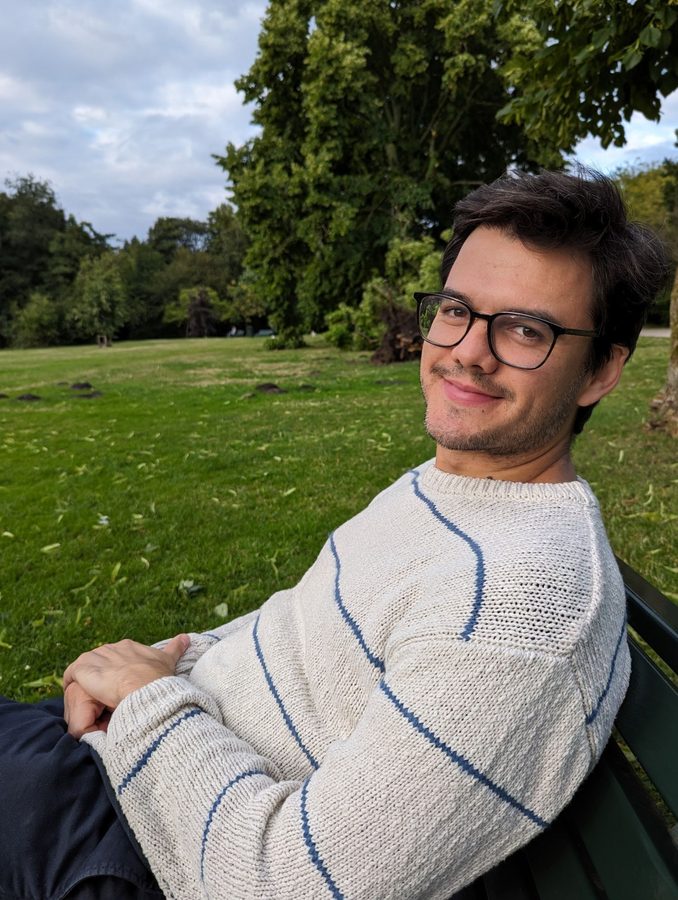 Daniel smiling at the camera, sitting on a park bench in a striped cream sweater.